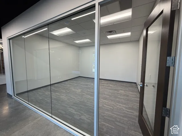Glass partition walls with an integrated wood-finish door, patterned carpet flooring, white walls, and a dropped ceiling with recessed lighting