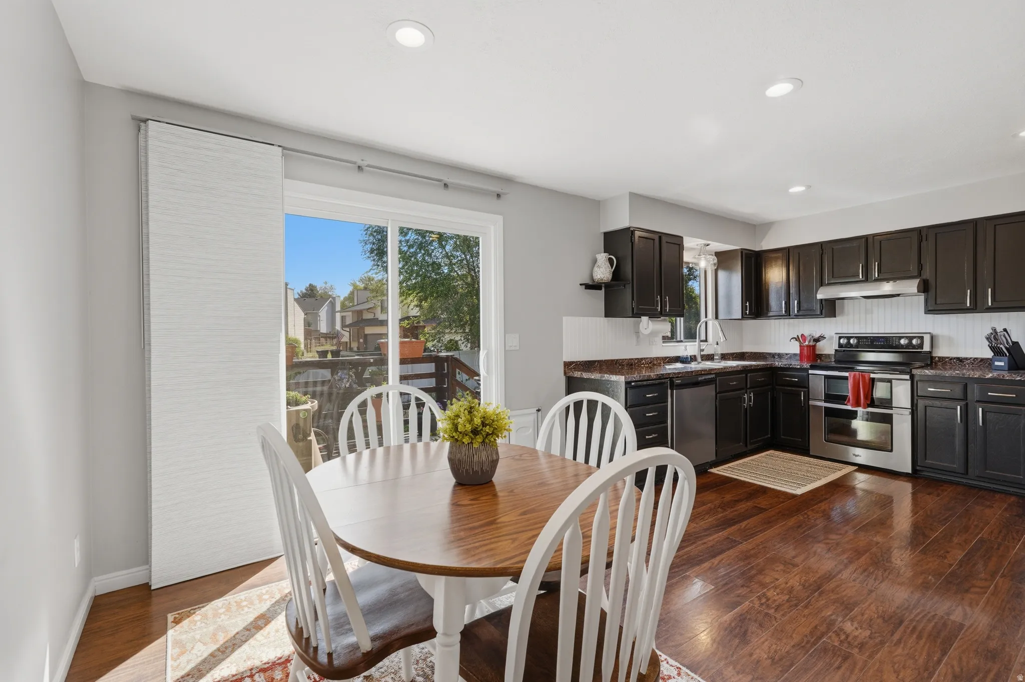 Kitchen and dining area featuring wood-finish flooring, dark cabinetry, stainless steel appliances, a white tile backsplash, and a sliding glass door