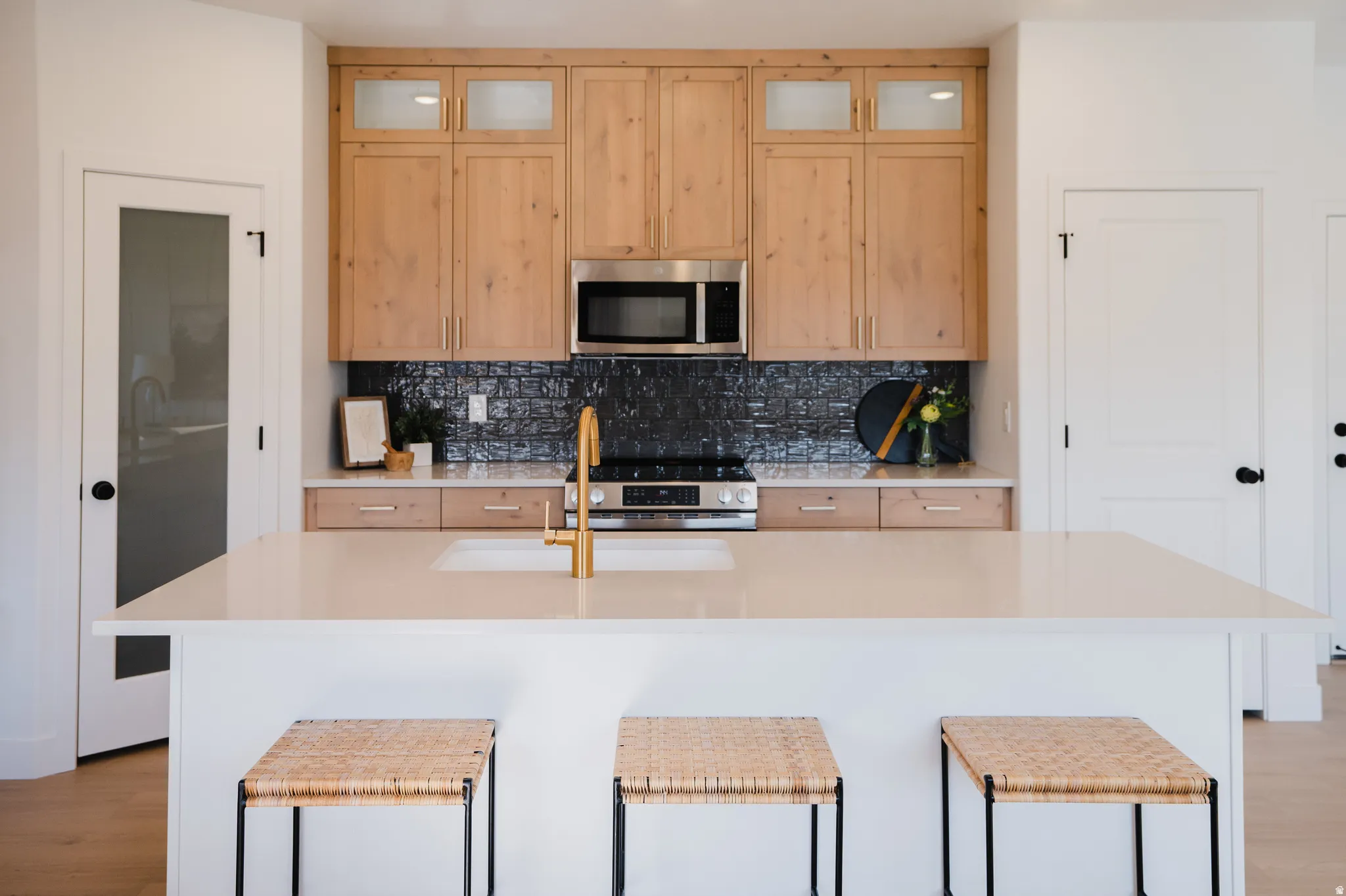 Kitchen featuring a large central island with a built-in sink and golden faucet, light wood-finish cabinetry, a textured black subway tile backsplash, stainless steel appliances, and quartz countertops