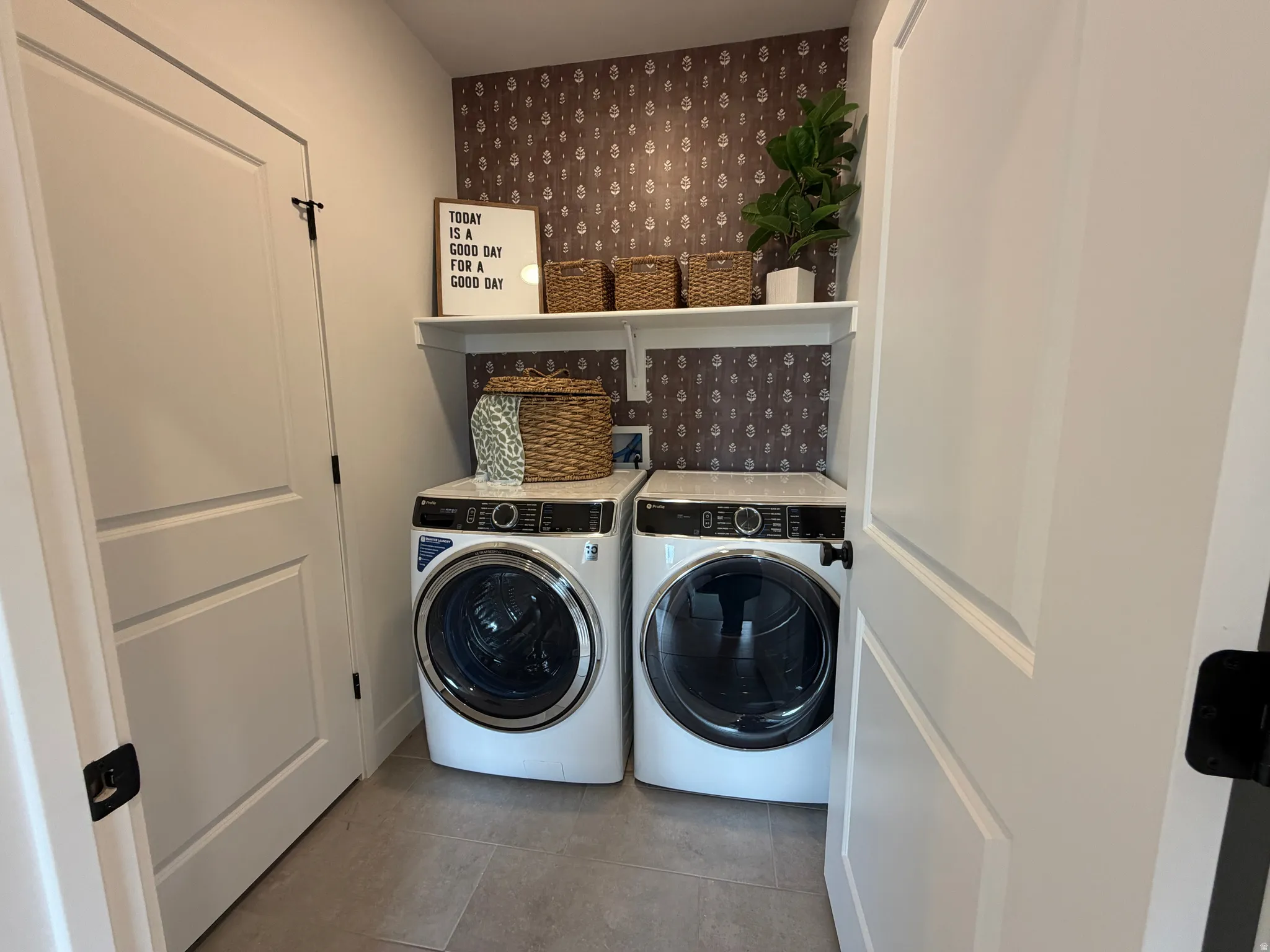 Dedicated laundry area featuring a patterned accent wall, light gray tile flooring, a built-in white shelf, and white panel doors with black hardware