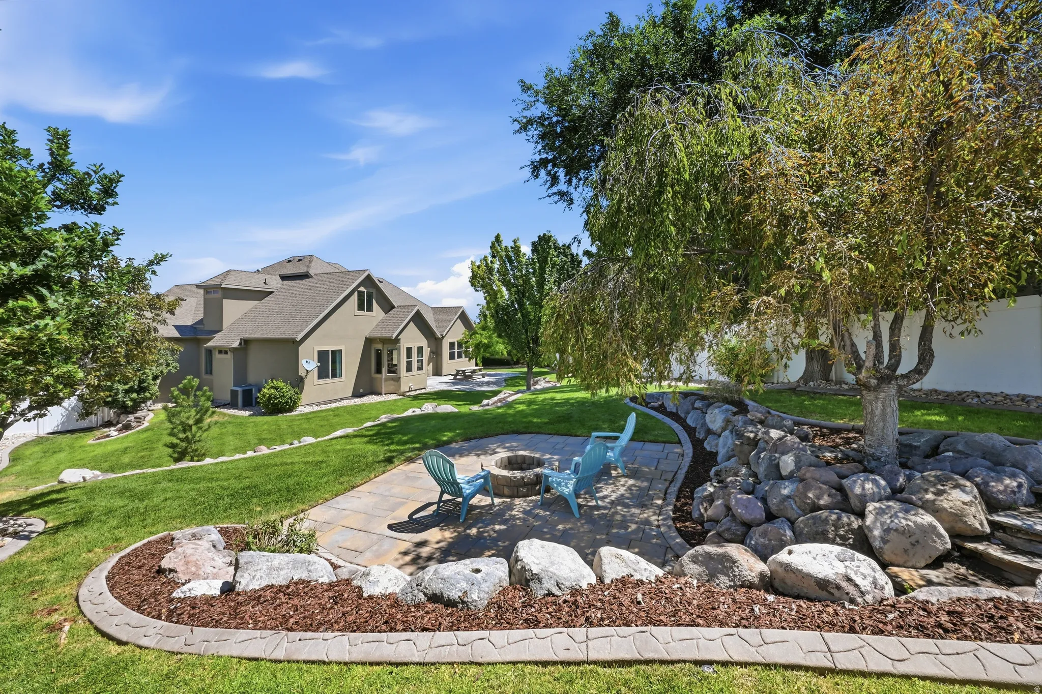 Expansive backyard featuring a stone patio with a built-in fire pit, mature trees, and natural rock landscaping