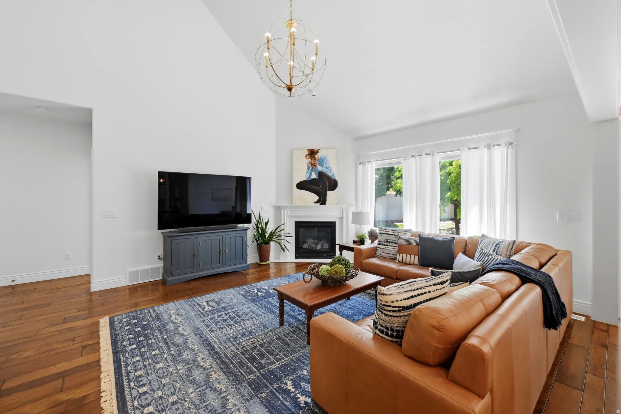 Living space featuring vaulted ceilings, wood-finish flooring, a decorative chandelier, a fireplace with a white mantel, and multiple windows providing natural light