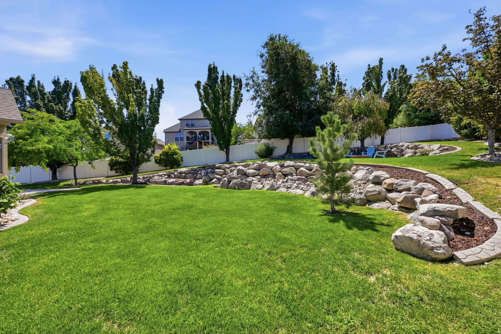 Expansive lawn area featuring a decorative rock wall, mature trees, a white perimeter fence, and a curved garden bed with decorative edging