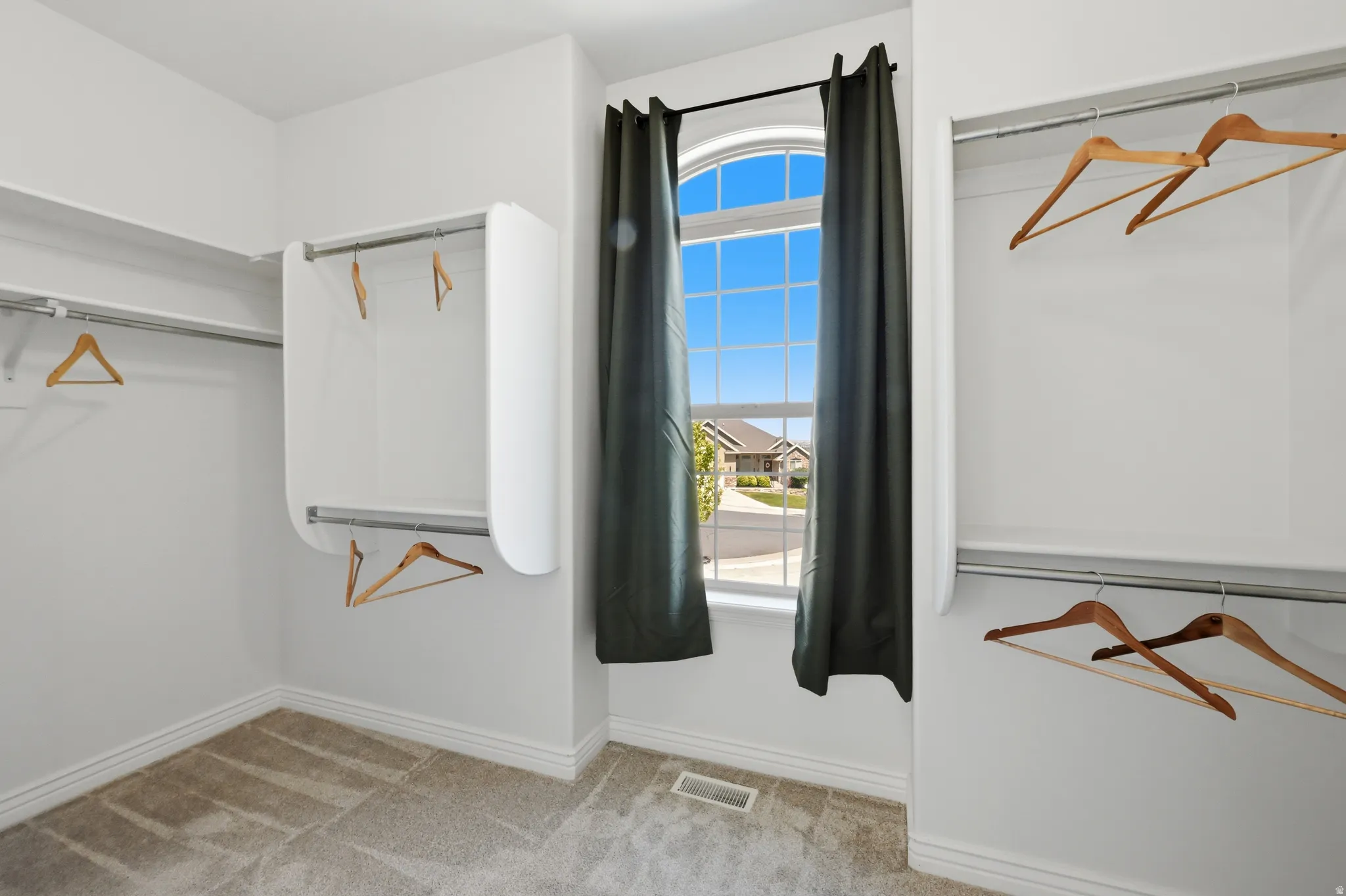 Walk-in closet featuring an arched window, light gray carpet flooring, white baseboards, and built-in shelving with hanging rods