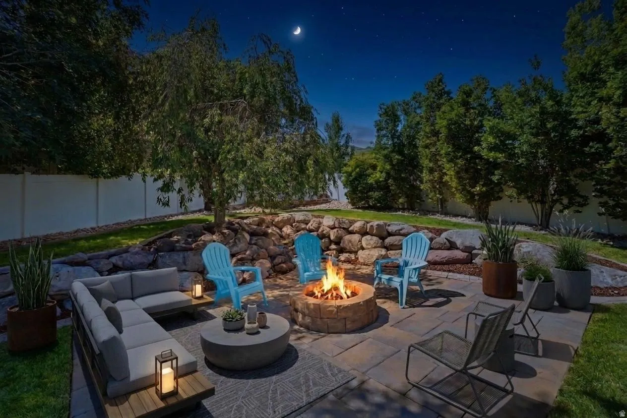 Paver patio featuring a stone-clad fire pit, tiered landscaping with natural rock retaining walls, mature trees, and a private perimeter fence