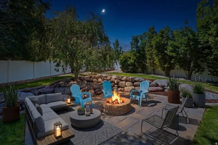 Paver patio featuring a stone-clad fire pit, tiered landscaping with natural rock retaining walls, mature trees, and a private perimeter fence
