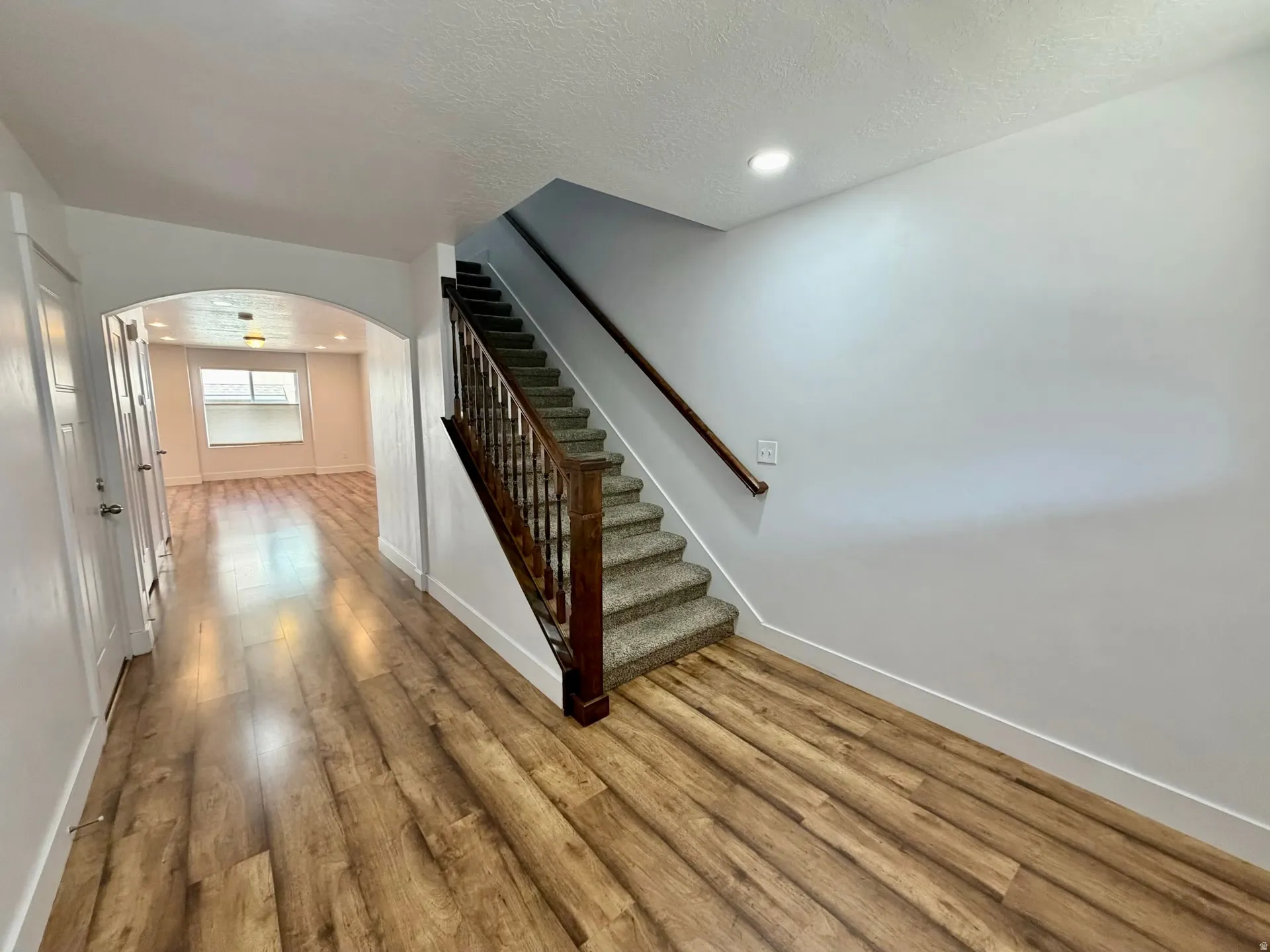 Foyer with wood-finish flooring, a carpeted staircase with a dark wood banister, and an arched doorway leading to a further room