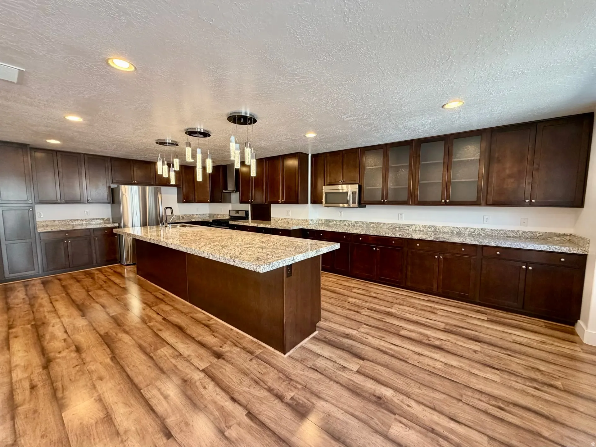 Kitchen featuring a large central island with a light-toned countertop, rich wood-finish cabinetry, wood-finish flooring, recessed lighting, and a contemporary chandelier