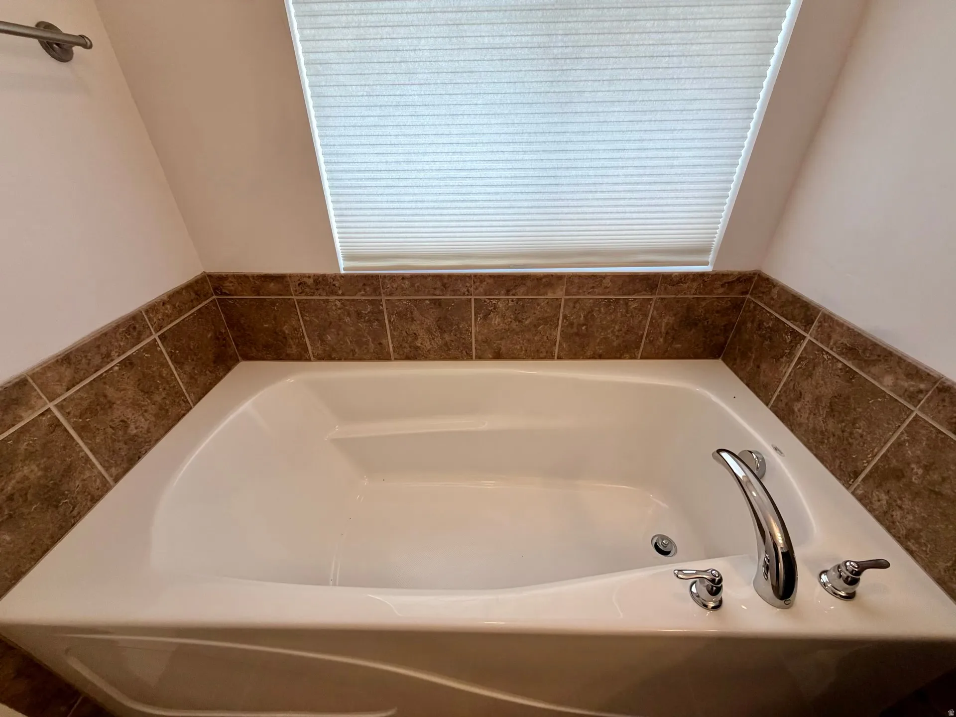 Built-in soaking tub with chrome fixtures, surrounded by brown tilework, set beneath a privacy blind