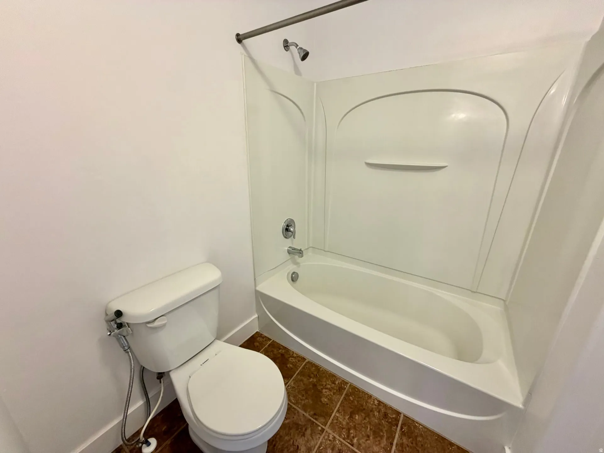 Bathroom featuring a white toilet, brown-toned tile flooring, and a shower-tub combination unit with a built-in shelf