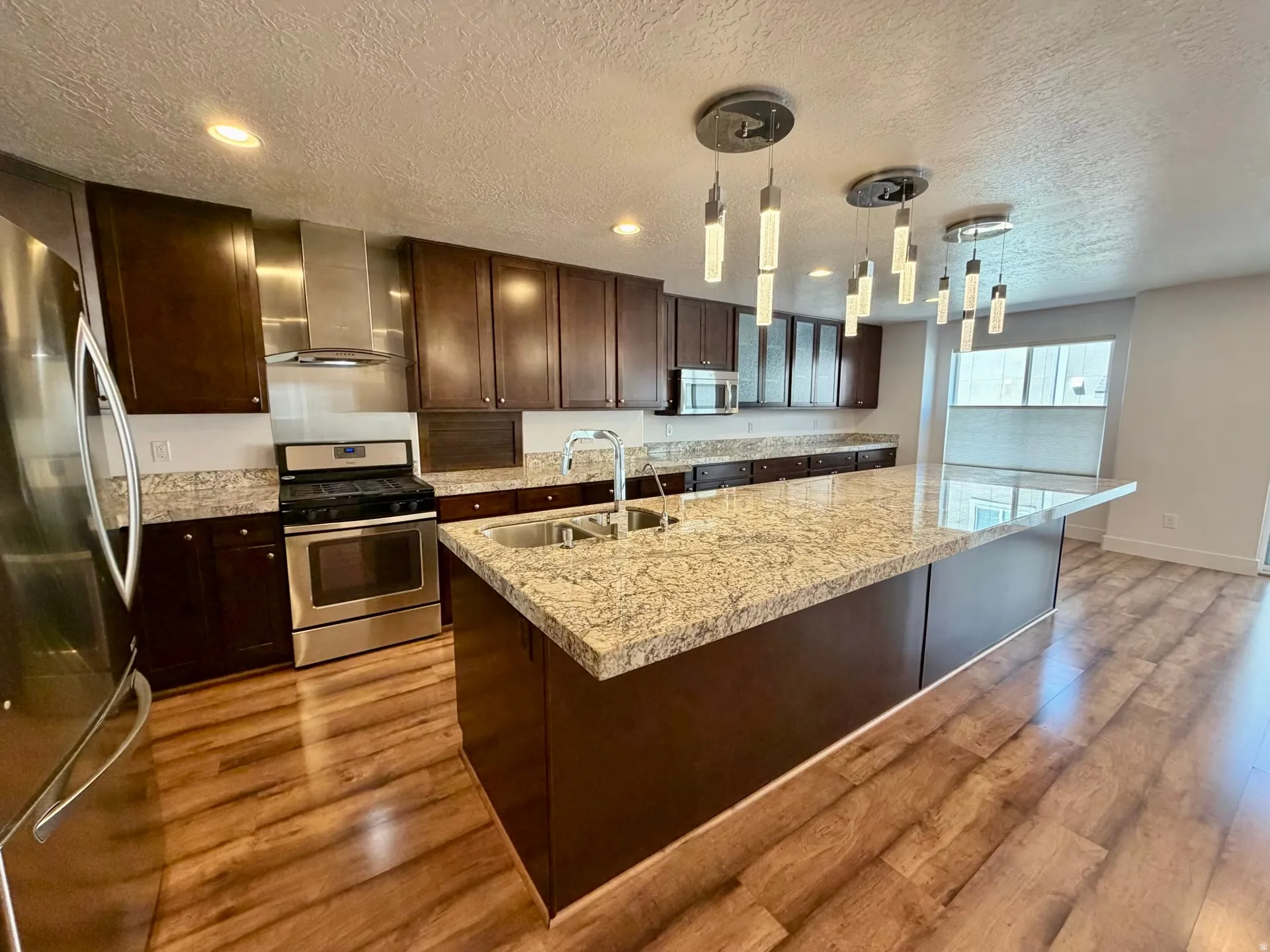 Spacious kitchen featuring an oversized island with a dual-basin stainless steel sink, granite-style countertops, dark wood cabinetry, stainless steel appliances, and wood-finish flooring