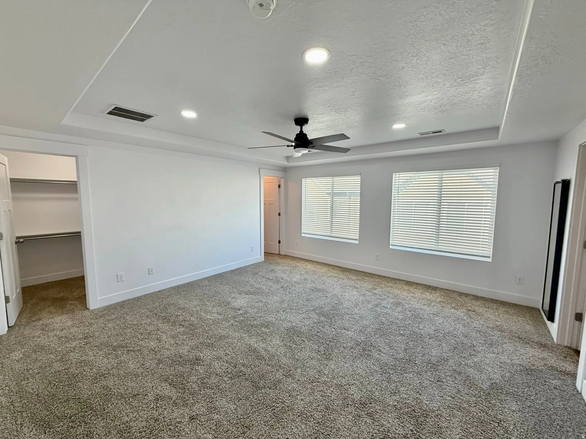 Spacious carpeted room featuring a tray ceiling with recessed lighting, a ceiling fan, and two windows with blinds