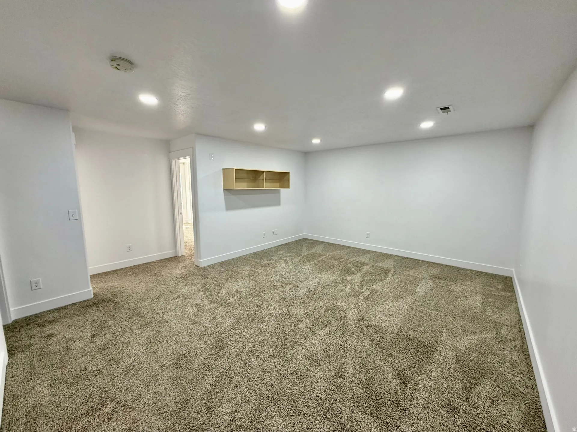 Spacious room featuring neutral-toned carpet, recessed lighting, a built-in light wood-finish shelving unit, and white baseboards