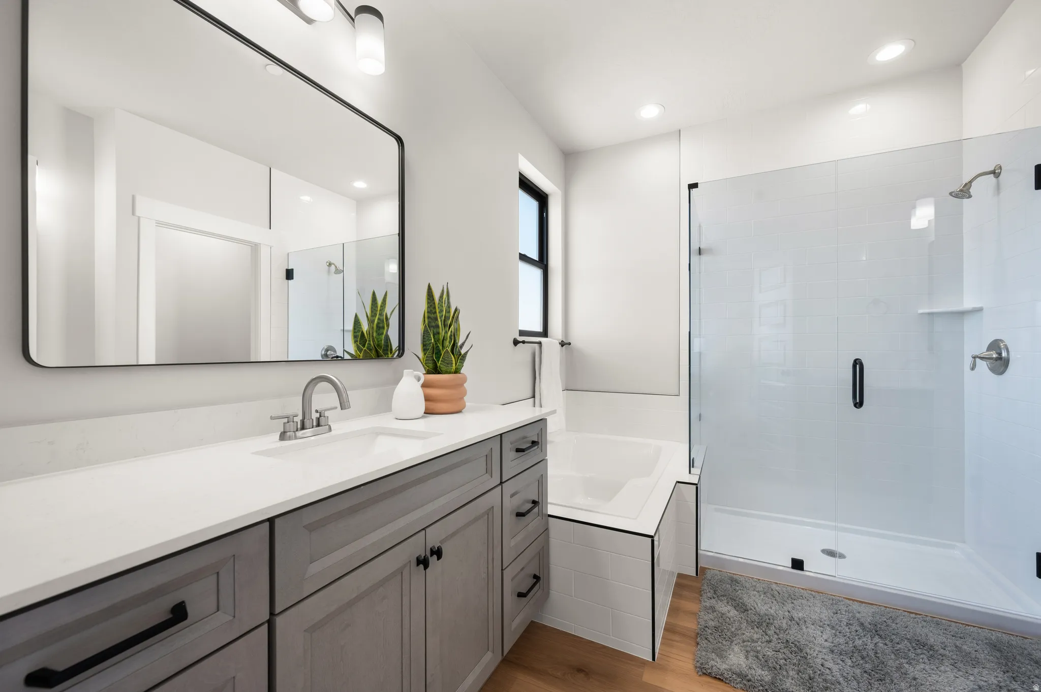 Contemporary bathroom featuring a spacious vanity with a white countertop and dual sinks, a large framed mirror, a separate shower with a clear glass enclosure, a built-in tub, and wood-finish flooring
