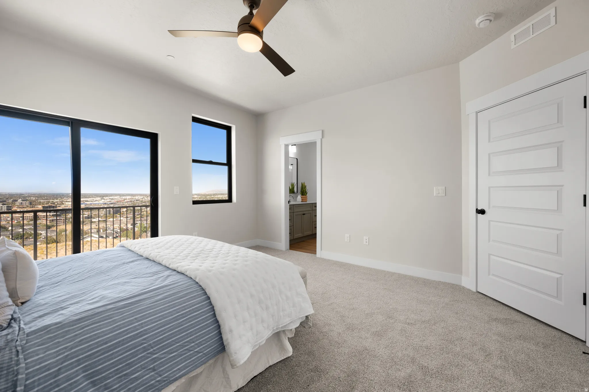 Spacious bedroom featuring light gray walls, plush carpeting, a multi-paneled white door with black hardware, a modern ceiling fan, and a floor-to-ceiling glass door opening to a balcony with metal railing