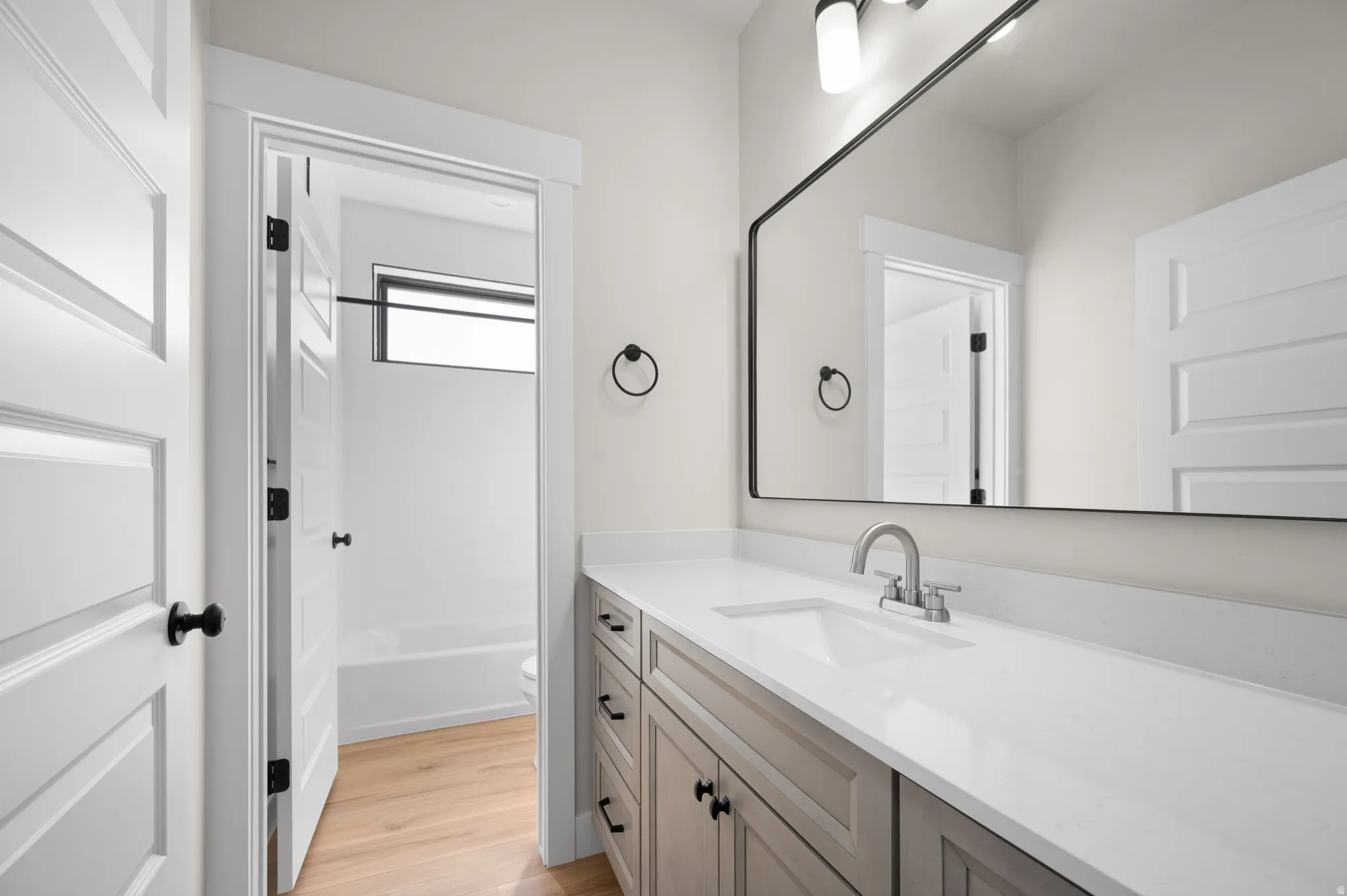 Bathroom featuring a light wood-finish floor, a rectangular undermount sink, a white countertop, a gray vanity with black hardware, and a large mirror with a black frame
