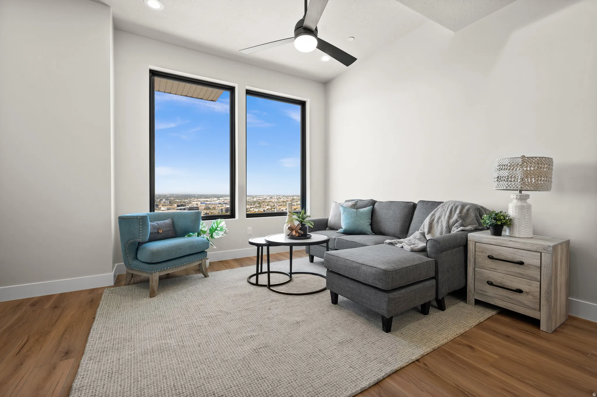 Two expansive windows frame the city skyline, complemented by a vaulted ceiling and a modern ceiling fan with integrated lighting