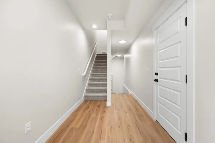 Contemporary hallway featuring wood-finish flooring, light grey painted walls, recessed lighting, and a five-panel interior door with matte black hardware