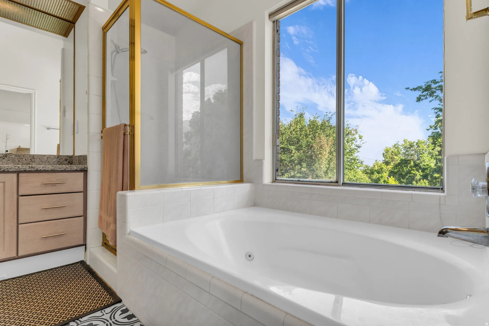 Deep soaking tub with a full-height window, white tile surround, and a gold-framed glass shower enclosure