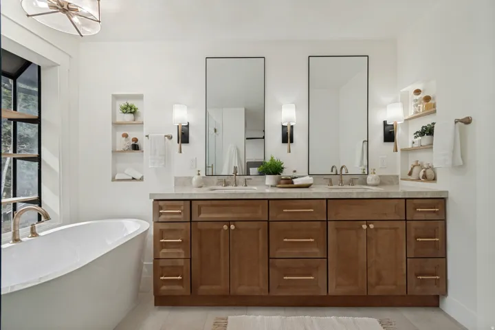Freestanding soaking tub with brushed gold fixture, double vanity with light wood-finish cabinetry and gold hardware, dual rectangular framed mirrors, and built-in wall shelving
