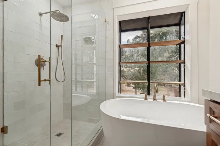 Freestanding soaking tub with brushed gold fixtures, a large window with horizontal wood-finish accents, and a glass-enclosed shower featuring subway tile and a rainfall showerhead with a handheld spray