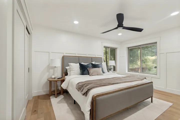 Bedroom with light wood-finish flooring, white paneled accent wall, and two large windows