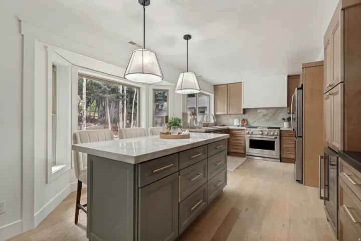 Kitchen featuring a large central island with a light-toned countertop, pendant lighting, built-in stainless steel appliances, light wood-finish flooring, and a bay window overlooking trees