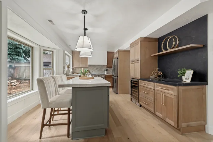 Kitchen featuring light wood-finish flooring, a large bay window, pendant lighting, a central island with a light-toned countertop, and a built-in wet bar with a dark stone-look backsplash and floating wood shelf