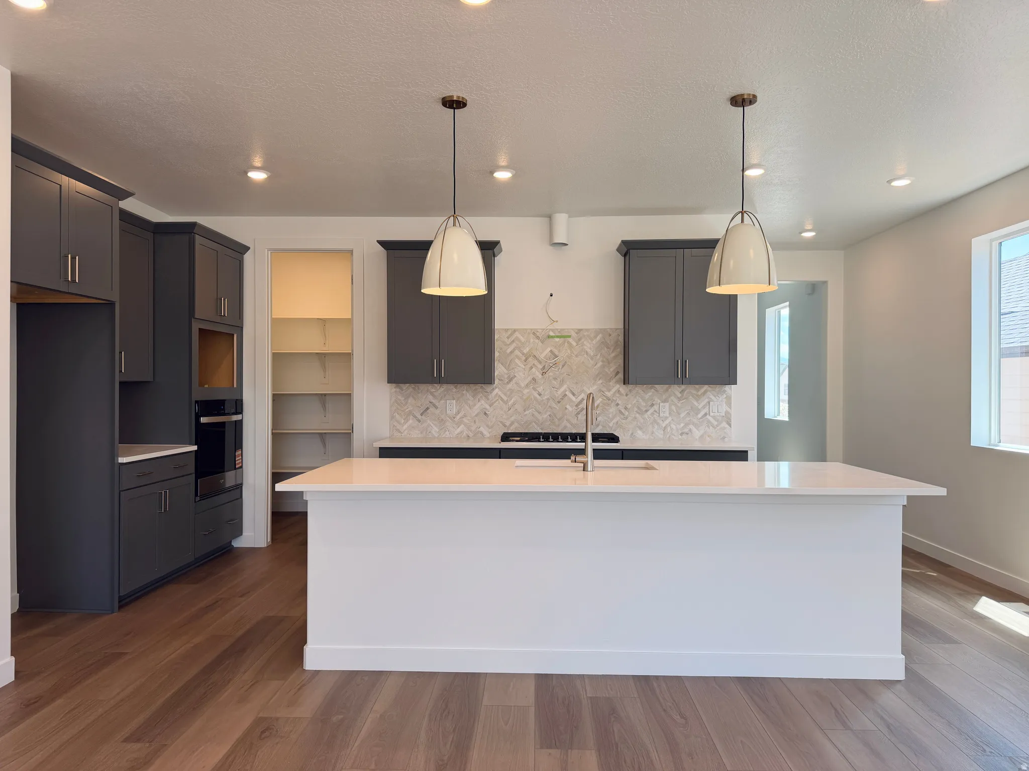 Spacious kitchen featuring an expansive central island, herringbone tile backsplash, dark wood-finish flooring, recessed lighting, and a pantry with built-in shelving