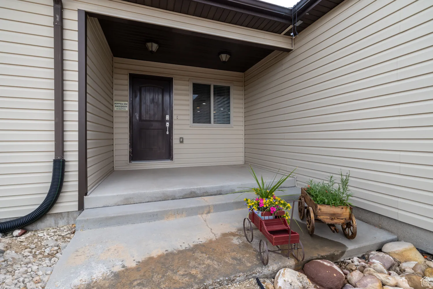 Covered concrete entry with a dark wood-finish door, light beige siding, and a single window with blinds