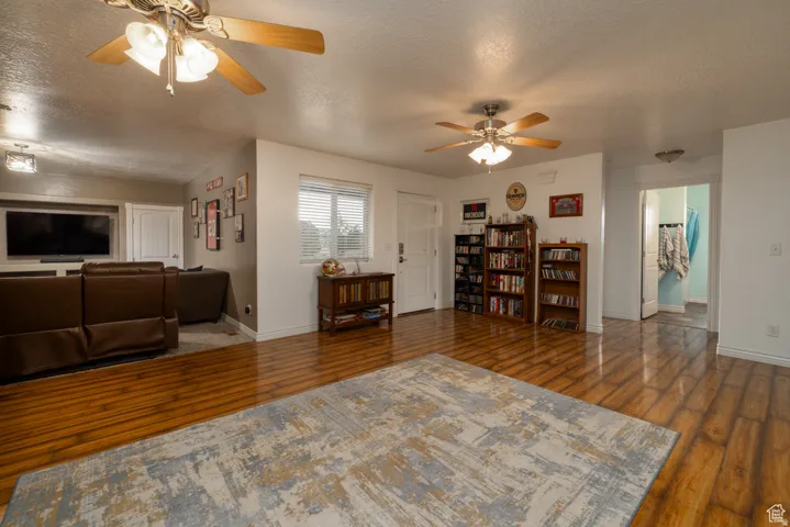 Spacious living area featuring wood-finish flooring, two ceiling fans with integrated lighting, and a prominent window with blinds