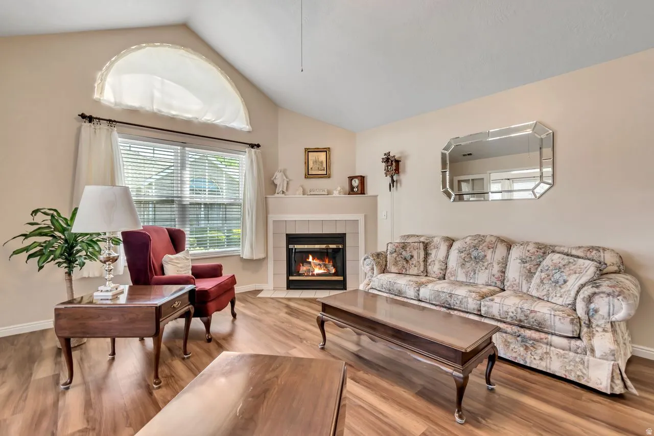 Living area featuring wood-finish flooring, a fireplace with a tiled surround, a vaulted ceiling, and a prominent window with an arched transom above