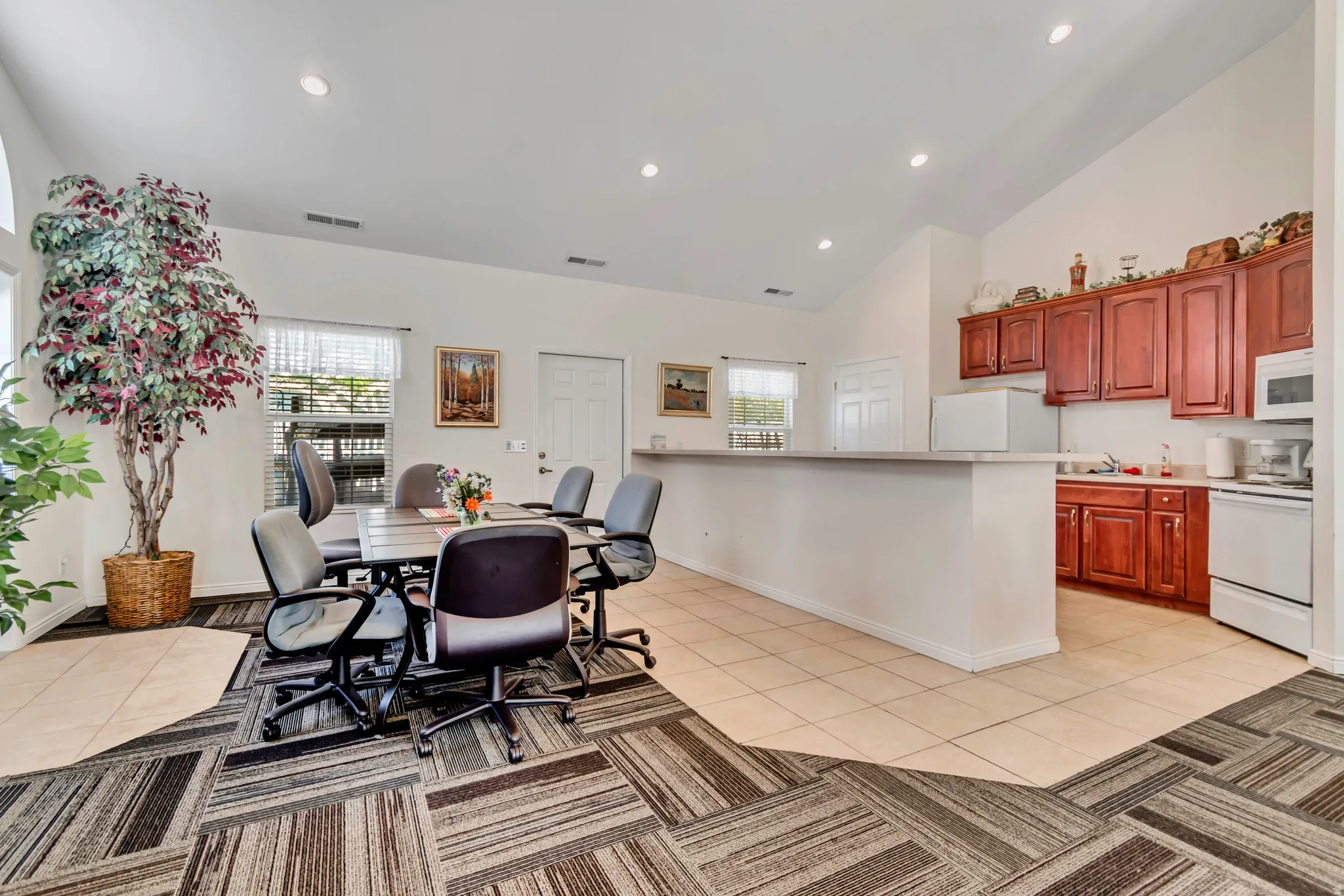 Open-concept common area featuring vaulted ceilings with recessed lighting, an integrated kitchenette with wood cabinetry and a pass-through counter, and a dining area with patterned carpet
