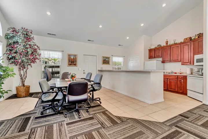 Open-concept common area featuring vaulted ceilings with recessed lighting, an integrated kitchenette with wood cabinetry and a pass-through counter, and a dining area with patterned carpet