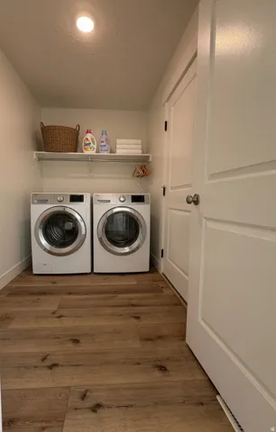 Dedicated laundry room featuring side-by-side front-load washers and dryers, wood-finish flooring, recessed ceiling lighting, open shelving with woven storage basket, and a full-height white six-panel entry door.  AI photographic version of the laundry ro
