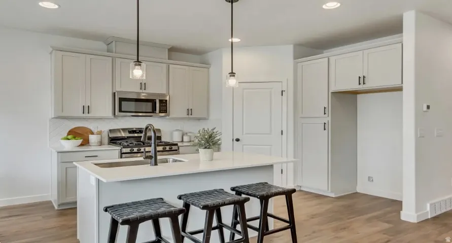 Kitchen featuring an island with a built-in sink, stainless steel appliances, light-toned cabinetry, a subway tile backsplash, and wood-finish flooring. Picture is of a similar home.