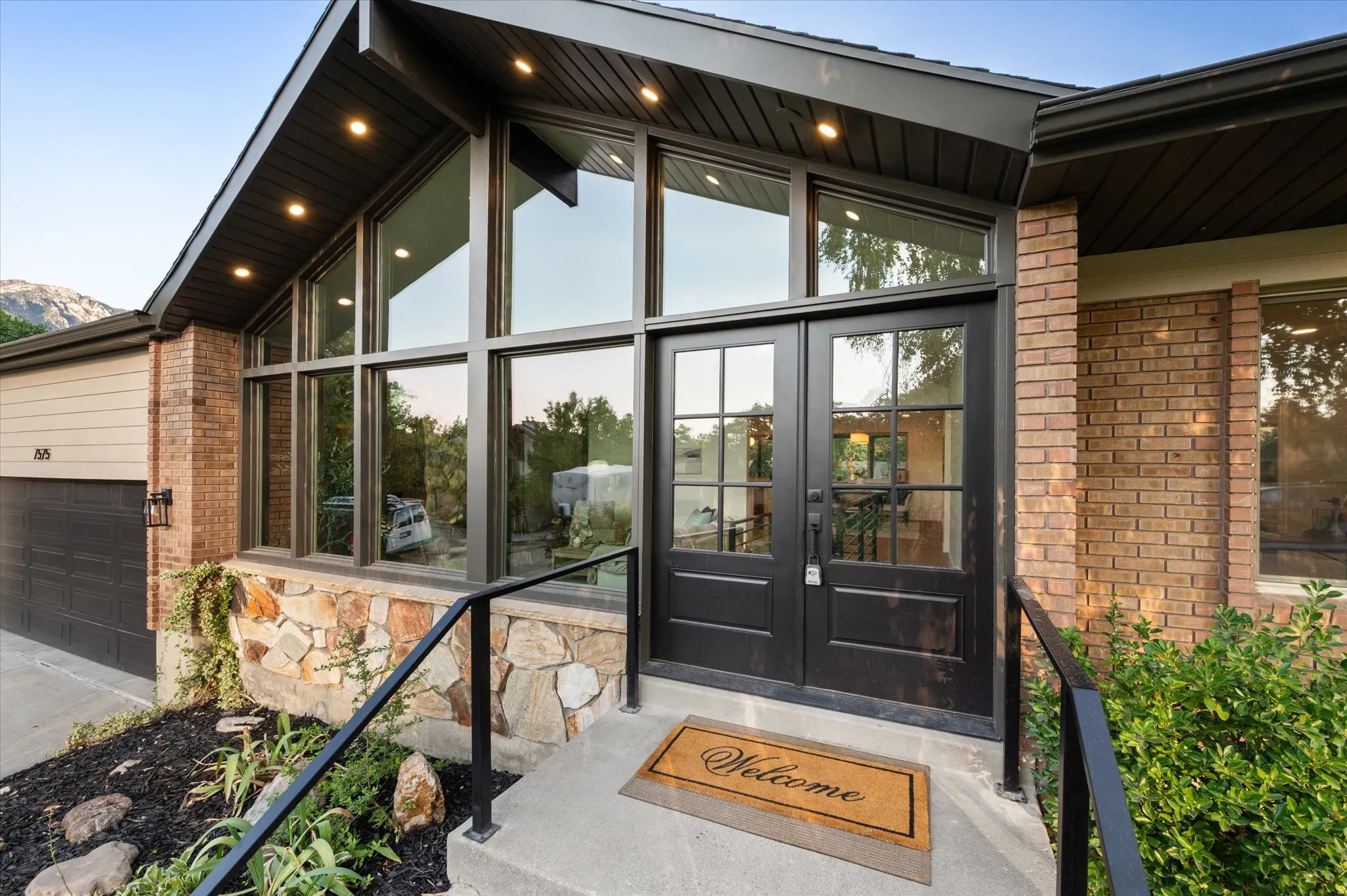 Exterior featuring a brick facade, stone accents, black-framed windows, double glass-paneled entry doors, and a concrete pathway with a black metal handrail