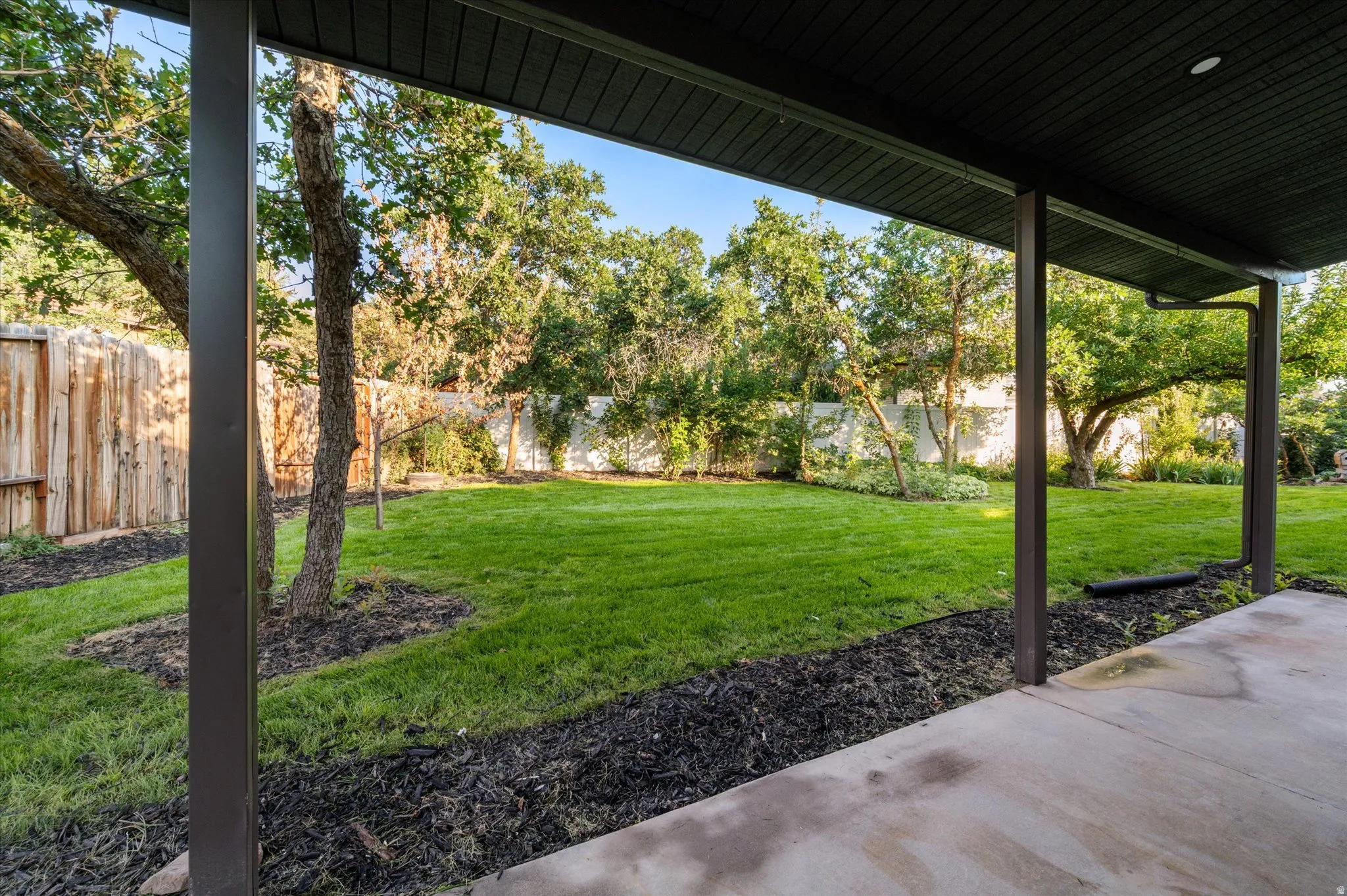 Covered concrete patio with dark support beams overlooking a spacious lawn with mature trees and wood fencing