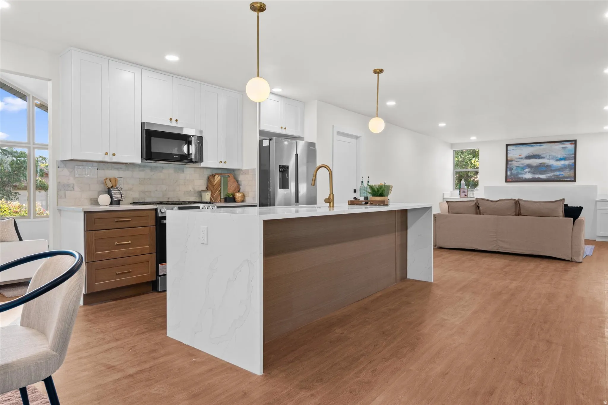 Open-concept kitchen featuring an island with a waterfall countertop, brass-finish fixtures, stainless steel appliances, white shaker cabinetry, and wood-finish flooring