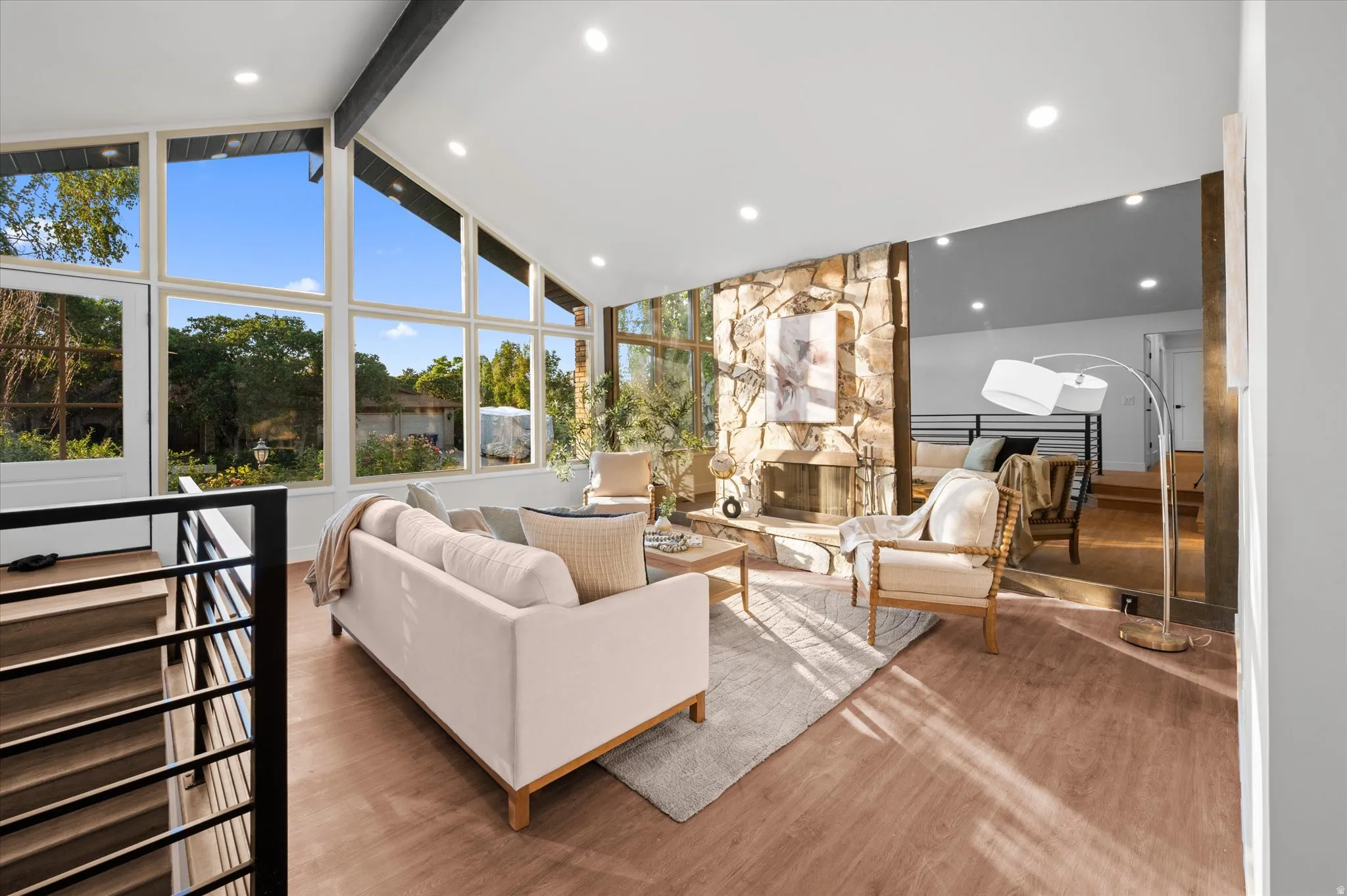 Vaulted ceilings with exposed beam, floor-to-ceiling windows, stone-clad fireplace, and wood-finish flooring