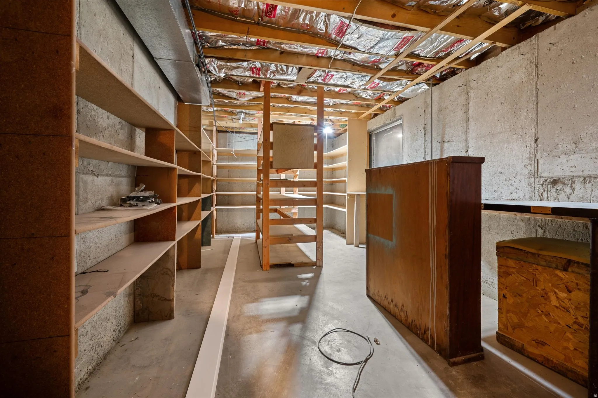 Unfinished basement space featuring concrete walls, exposed ceiling joists with insulation, and extensive built-in shelving