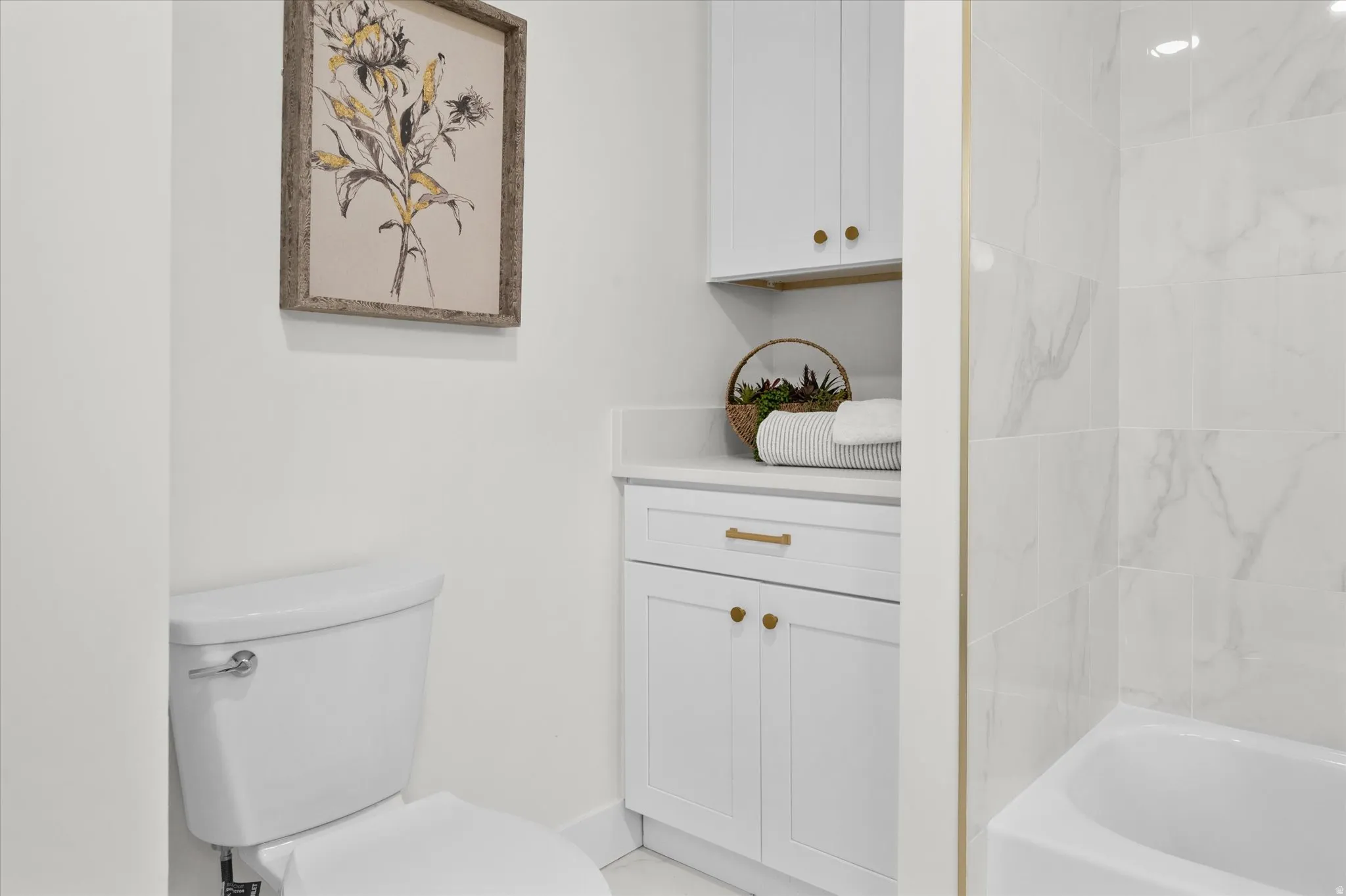 Bathroom featuring a white toilet, built-in white cabinetry with gold-finish hardware, a white countertop, and white tiled walls surrounding a bathtub