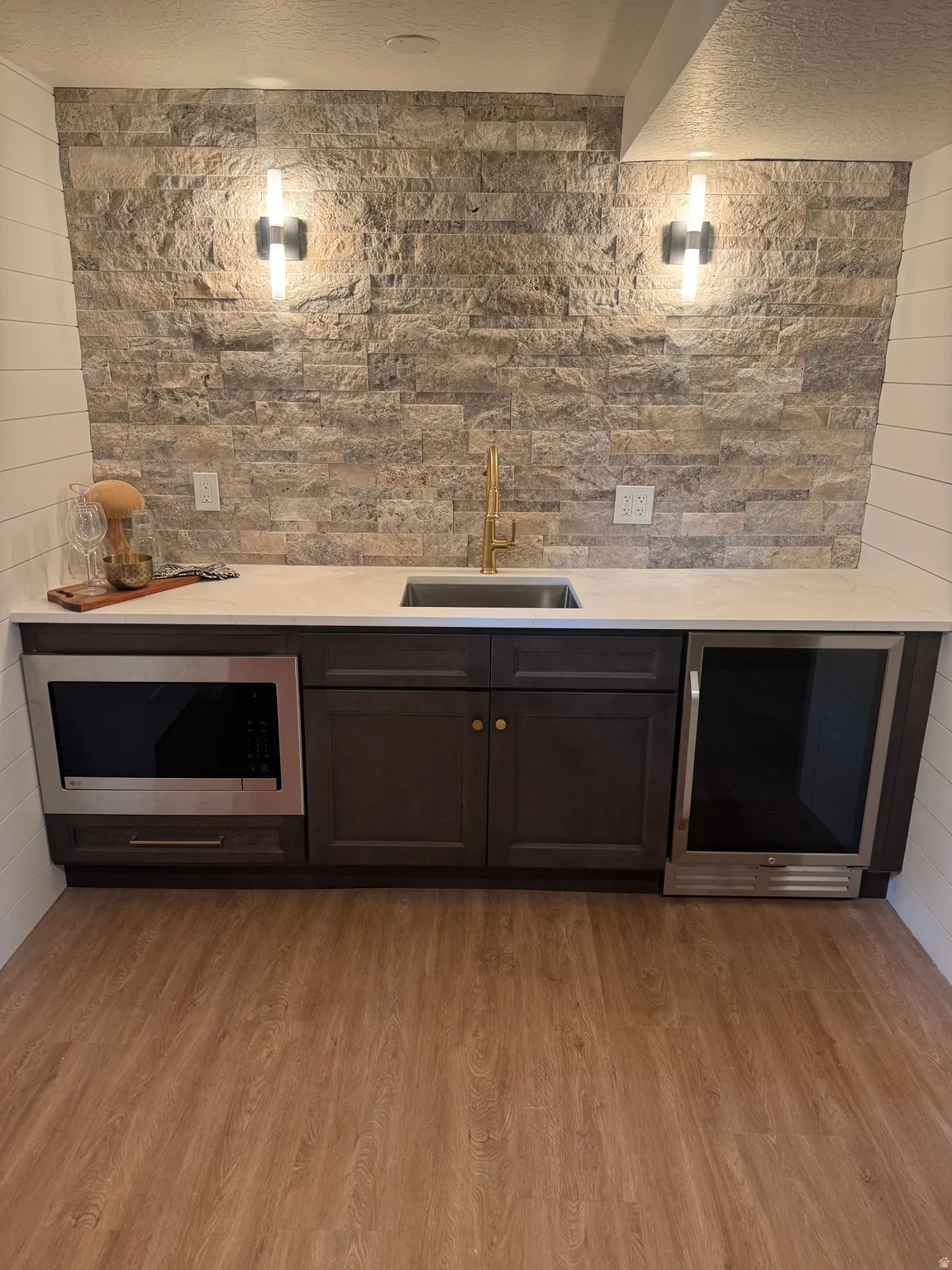 Wet bar featuring stainless steel appliances, including a built-in microwave and beverage cooler, dark cabinetry with brass hardware, white quartz countertop, brushed brass faucet, and natural stone tile backsplash with integrated sconce lighting