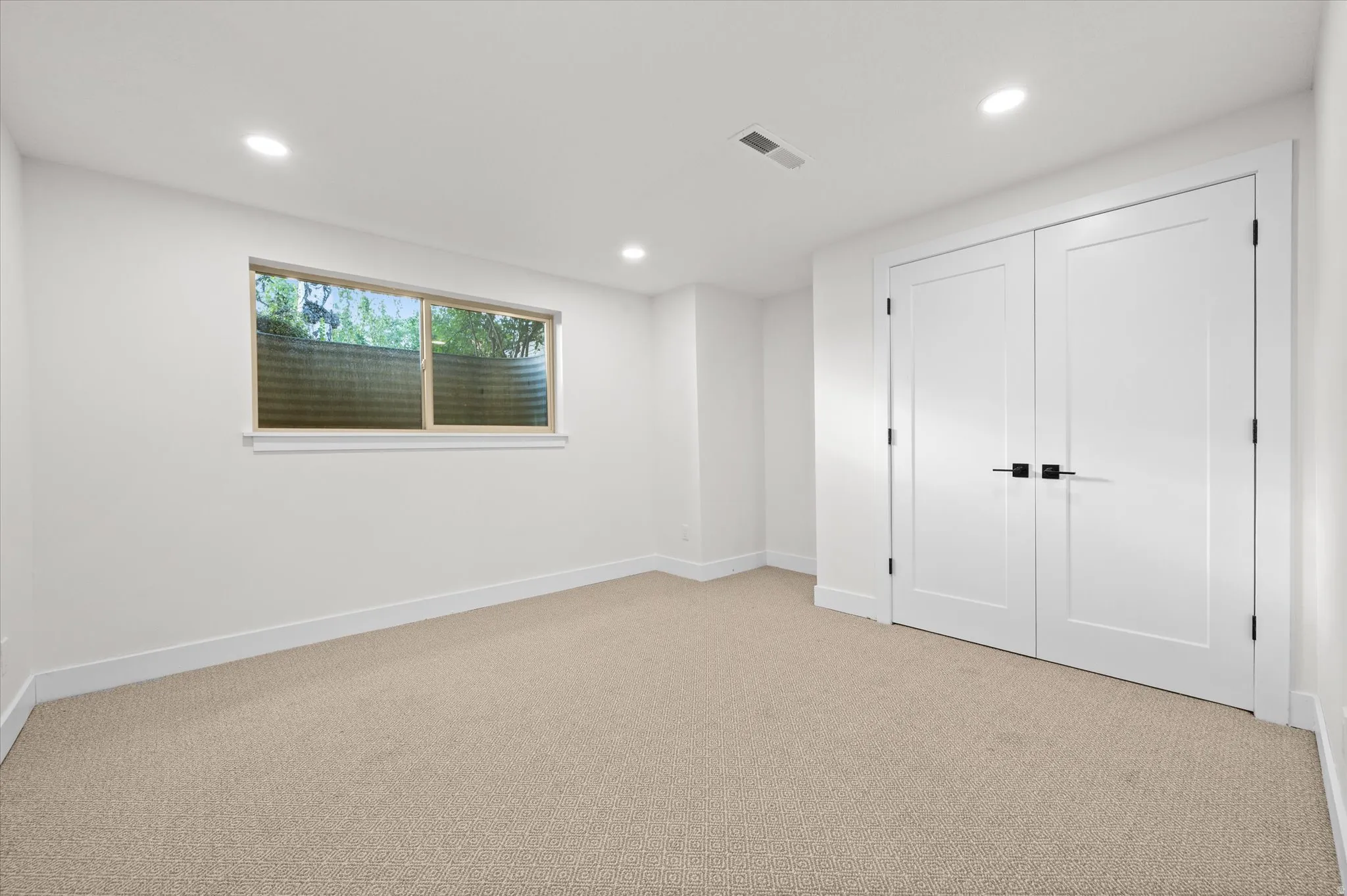 Carpeted room featuring a window with a window well, recessed lighting, and a pair of panel doors with matte black hardware