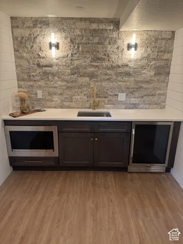 Wet bar featuring stainless steel appliances, including a built-in microwave and beverage cooler, dark cabinetry with brass hardware, white quartz countertop, brushed brass faucet, and natural stone tile backsplash with integrated sconce lighting