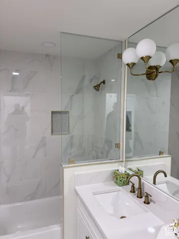 Sleek bathroom featuring a built-in tub with a glass shower panel, white marble-style wall tiling, and a vanity with an integrated sink