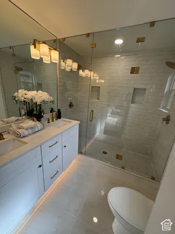 Dual vanity with undermount sinks, frameless glass shower enclosure, large format floor tiling, recessed shower shelving, and gold-finish fixtures