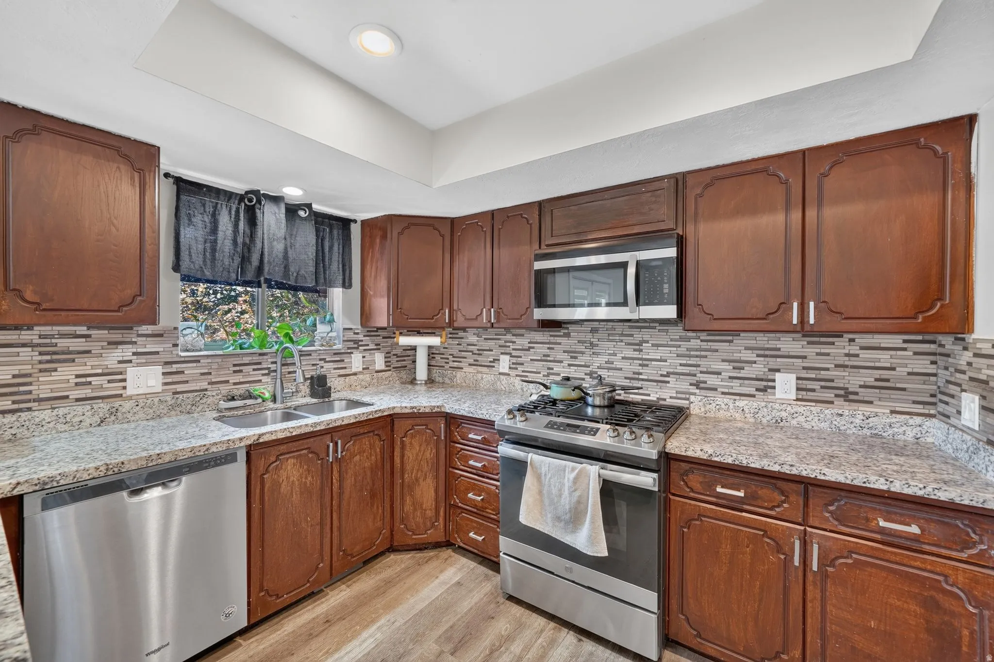 Kitchen featuring wood cabinetry, stainless steel appliances, a double basin sink, light-colored countertops, and a tiled backsplash