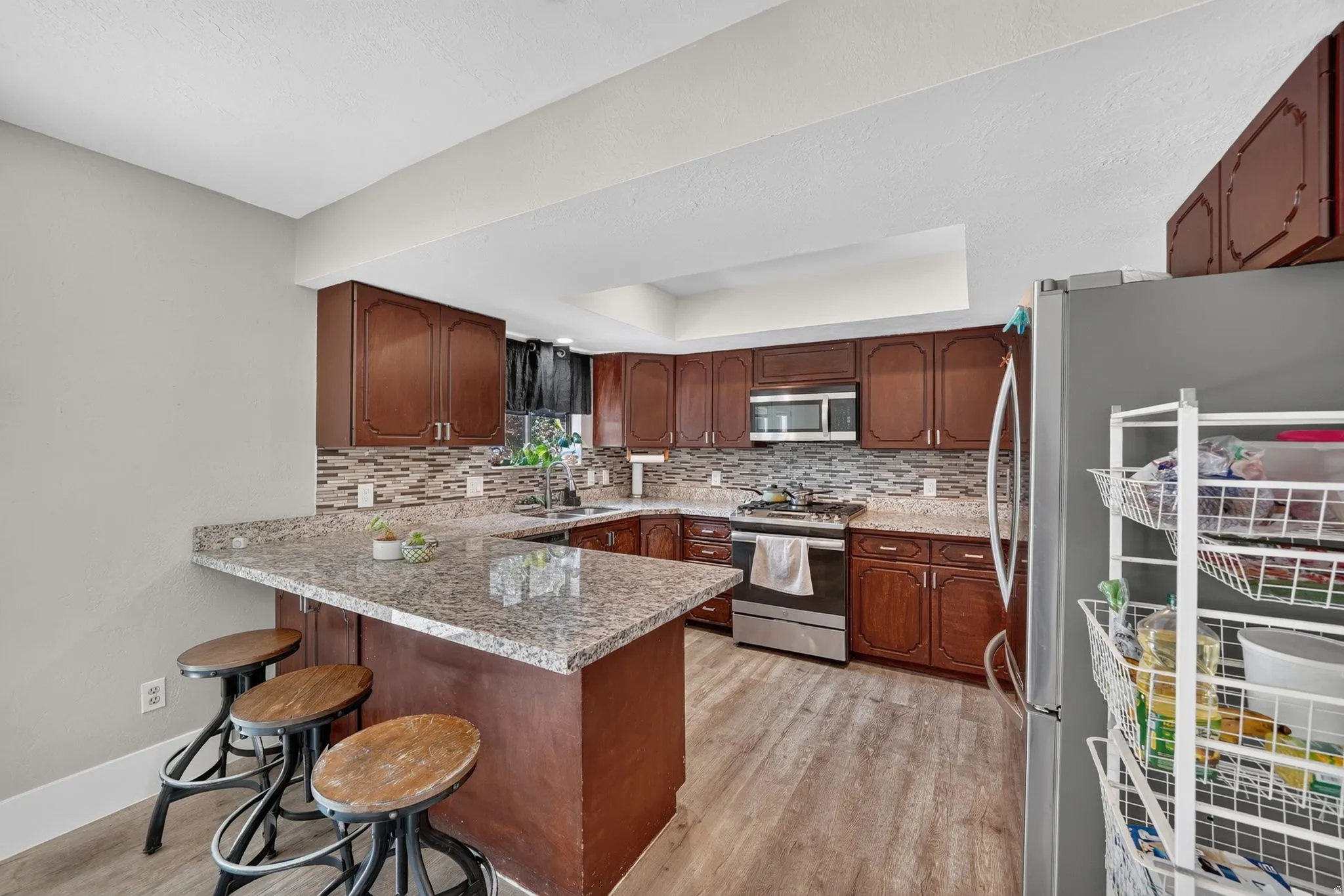 Kitchen featuring wood-finish cabinetry, granite-style countertops, stainless steel appliances, a mosaic tile backsplash, and wood-finish flooring