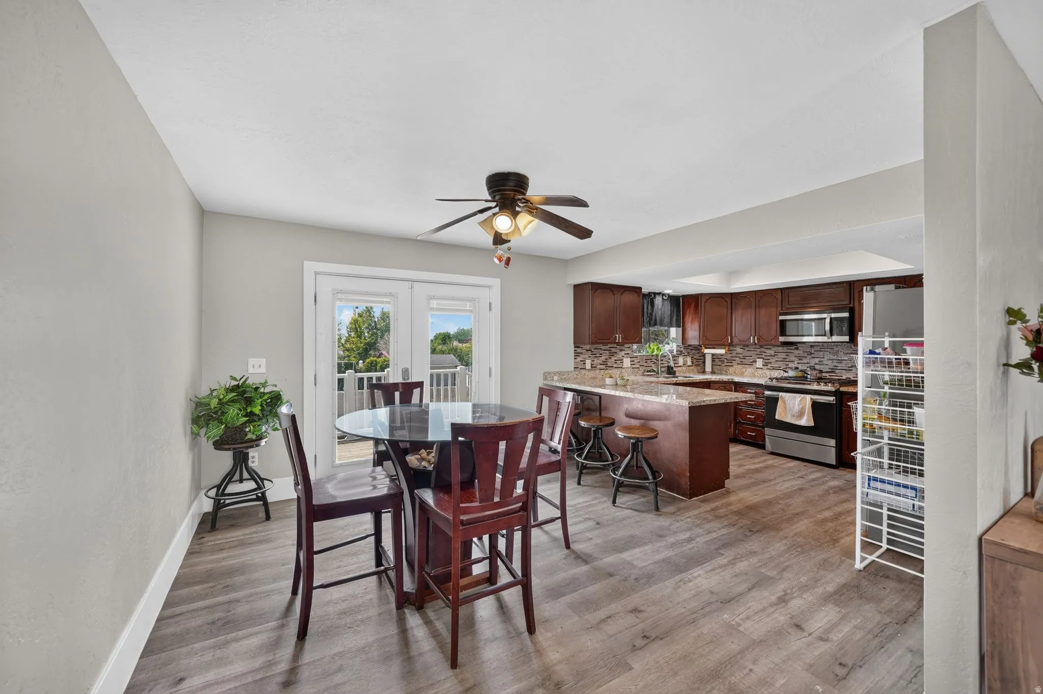 Open-concept living and kitchen space featuring wood-finish flooring throughout, rich cabinetry with integrated appliances, a ceiling fan with light fixture, and glass sliding doors opening to an outdoor deck